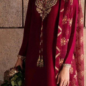 May include: A burgundy-colored ensemble featuring intricate gold embroidery. The long-sleeved tunic has a keyhole neckline and is paired with a matching scarf. The scarf has a floral pattern. The outfit is accessorized with gold jewelry and tassels.