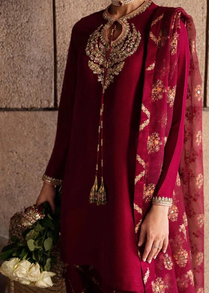 May include: A burgundy-colored ensemble featuring intricate gold embroidery. The long-sleeved tunic has a keyhole neckline and is paired with a matching scarf. The scarf has a floral pattern. The outfit is accessorized with gold jewelry and tassels.