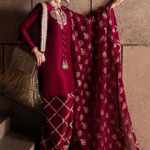 May include: Burgundy-colored formal ensemble featuring a long-sleeved tunic with intricate gold embroidery around the neckline and cuffs. The outfit includes matching pants with a diamond pattern and a sheer, embellished shawl. A gold fringed handbag completes the look.