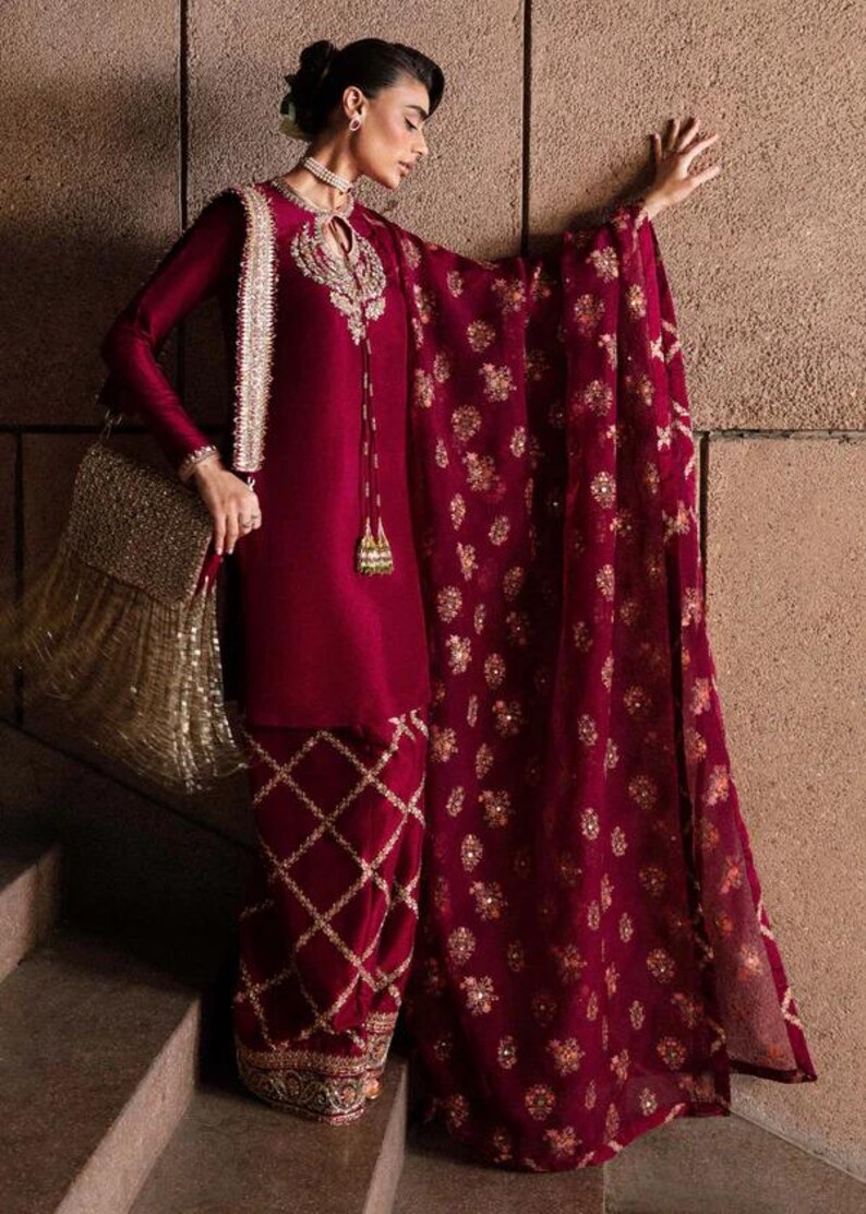 May include: Burgundy-colored formal ensemble featuring a long-sleeved tunic with intricate gold embroidery around the neckline and cuffs. The outfit includes matching pants with a diamond pattern and a sheer, embellished shawl. A gold fringed handbag completes the look.