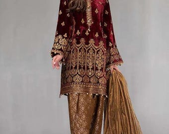May include: Burgundy velvet tunic and matching pants with gold embroidery. The long-sleeved tunic features intricate gold detailing around the neckline, cuffs, and hem. The pants have a matching gold pattern. A gold scarf is draped over the arm.