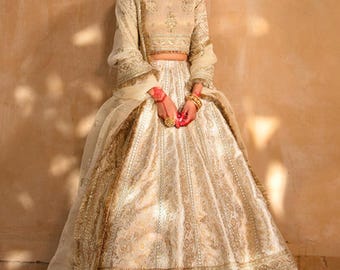 May include: A cream-colored, long-sleeved formal dress with intricate gold embroidery. The dress features a fitted bodice and a full skirt. The sleeves are long and flowing, and the dress is adorned with gold embellishments. The outfit is a traditional style.