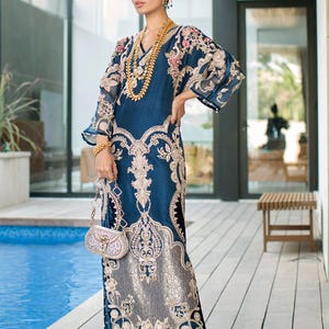 May include: A long, dark blue dress with intricate gold embroidery and embellishments. The dress features a V-neck, bell sleeves, and a flowing skirt. The woman is wearing gold jewelry and carrying a small, beaded purse. The background includes a pool and wooden deck.