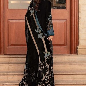 May include: A long, black velvet garment with intricate floral embroidery in teal and cream. The garment features a flowing design with a detailed border and accents on the sleeves. The elegant piece is suitable for special occasions.