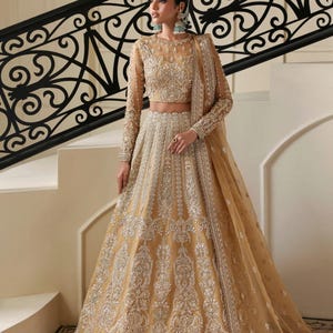 May include: A golden-yellow formal gown with intricate silver embroidery. The long-sleeved top and full skirt are embellished with detailed patterns. A matching sheer shawl drapes over the shoulder. The gown is worn in front of a staircase.