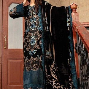 May include: A black velvet dress with intricate gold and teal floral embroidery. The dress features long sleeves and a matching shawl. The hemline and shawl edges are trimmed with teal lace. The woman is wearing teal heels.