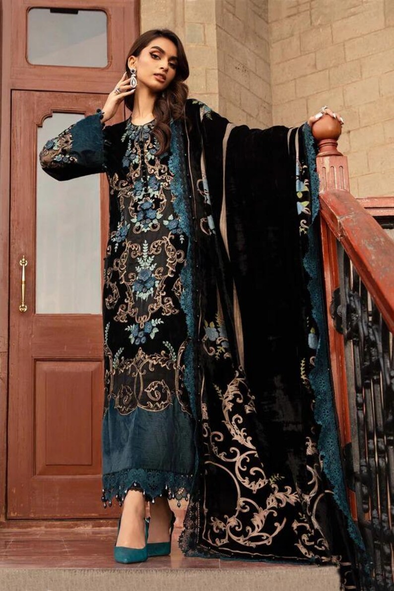 May include: A black velvet dress with intricate gold and teal floral embroidery. The dress features long sleeves and a matching shawl. The hemline and shawl edges are trimmed with teal lace. The woman is wearing teal heels.
