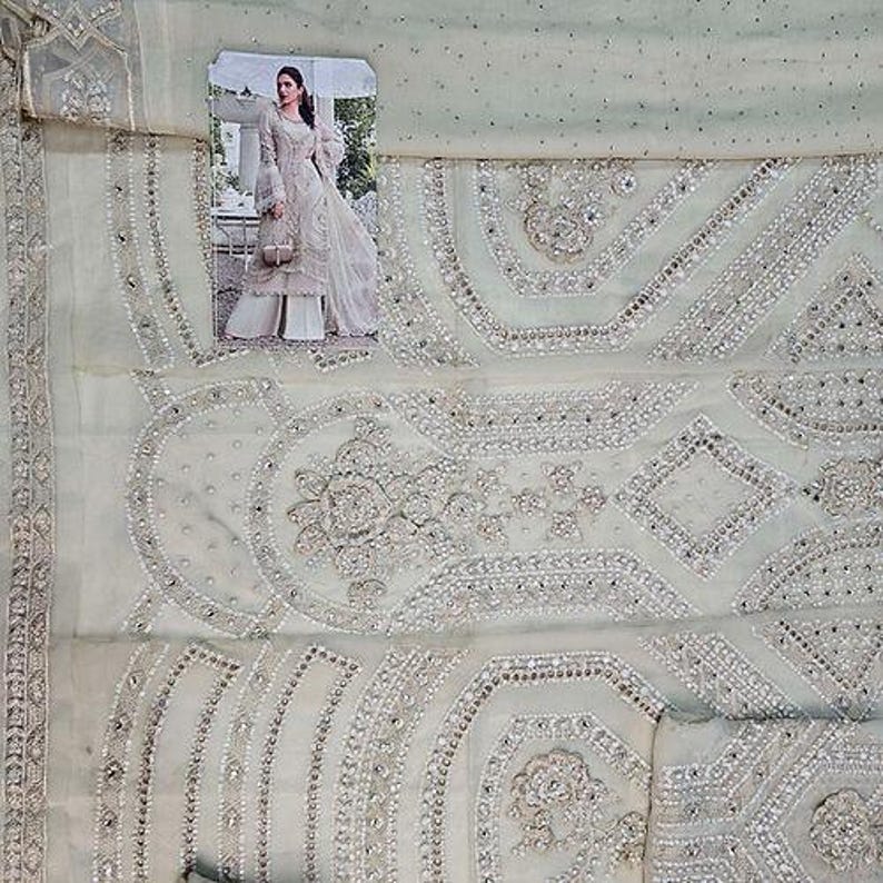 May include: A light green fabric with intricate embroidery and embellishments. The design features floral and geometric patterns, with a small image of a woman in a white dress. The fabric appears to be a delicate material, possibly for a formal outfit.