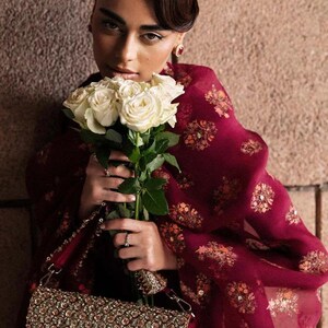 May include: A woman holding a bouquet of white roses, wearing a burgundy shawl with floral patterns. She is also holding a beaded handbag with a silver chain. The background is a textured, neutral-toned wall.