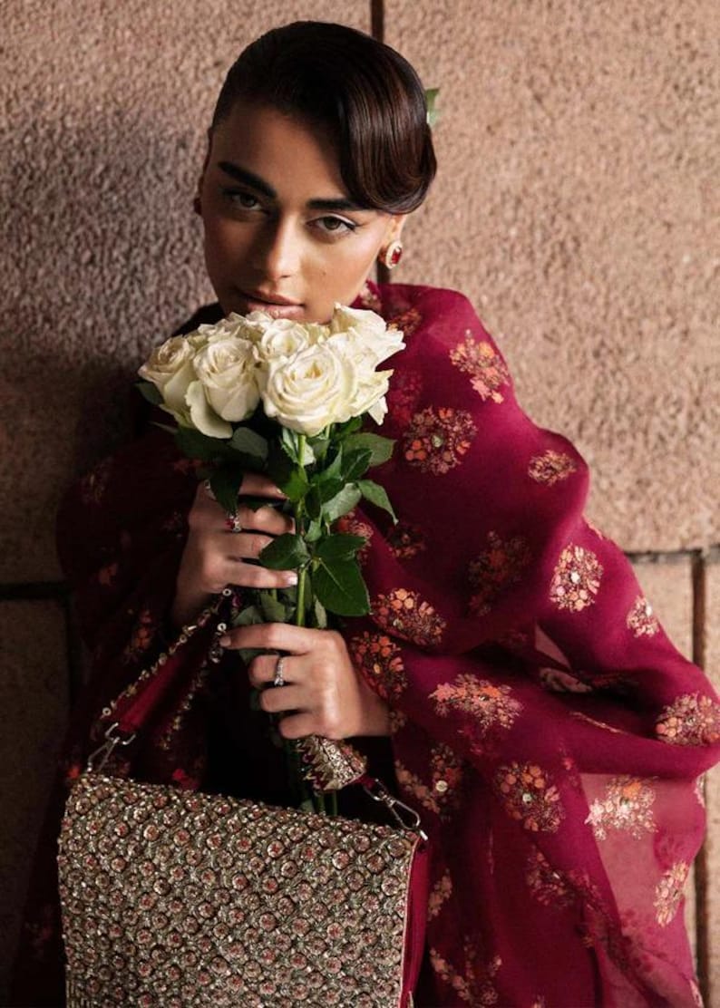 May include: A woman holding a bouquet of white roses, wearing a burgundy shawl with floral patterns. She is also holding a beaded handbag with a silver chain. The background is a textured, neutral-toned wall.
