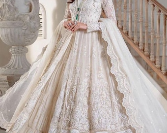 May include: A long, ivory-colored gown with intricate lace detailing. The dress features long sleeves, a flowing skirt, and a matching shawl. The background includes a staircase and decorative elements, suggesting a formal occasion.