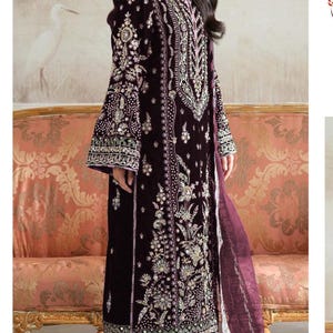 May include: A long, dark burgundy velvet dress with intricate floral embroidery and embellishments. The dress features long sleeves with flared cuffs and a matching burgundy skirt. A sheer burgundy scarf drapes from the shoulder. The dress is a formal wear garment.