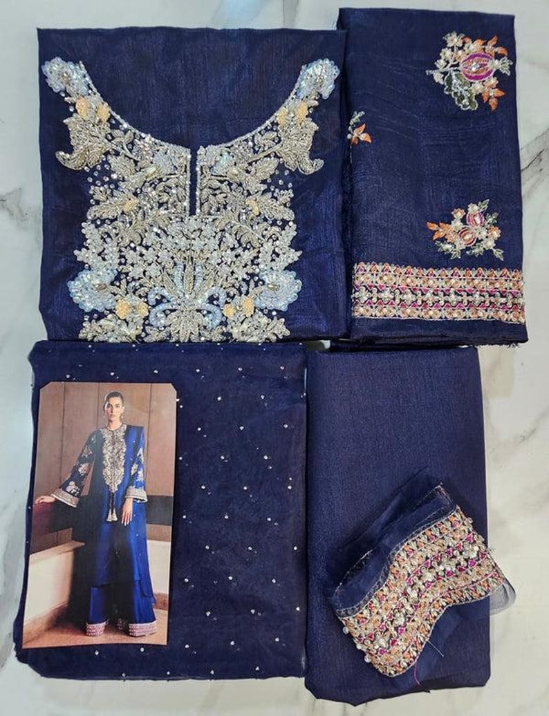 Puede incluir: Conjunto bordado azul marino. La parte superior presenta intrincados adornos plateados y dorados alrededor del escote. El conjunto incluye piezas de tela a juego, una con una foto de una persona que lleva el conjunto y un ribete decorativo.