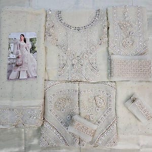 May include: An off-white, intricately embroidered formal outfit. The ensemble includes a top, pants, and a scarf, all adorned with detailed silver embellishments. A small picture of a woman in a similar outfit is included.