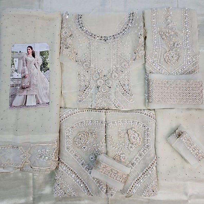 May include: An off-white, intricately embroidered formal outfit. The ensemble includes a top, pants, and a scarf, all adorned with detailed silver embellishments. A small picture of a woman in a similar outfit is included.