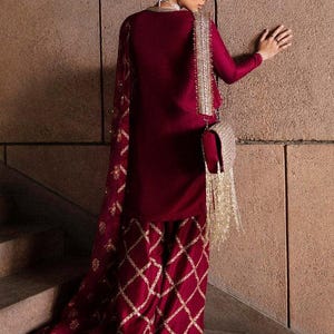 May include: A burgundy ensemble featuring a long-sleeved top and matching pants with a diamond pattern. The outfit includes a sheer scarf and a small, embellished purse with gold fringe. The pants and purse have gold accents.