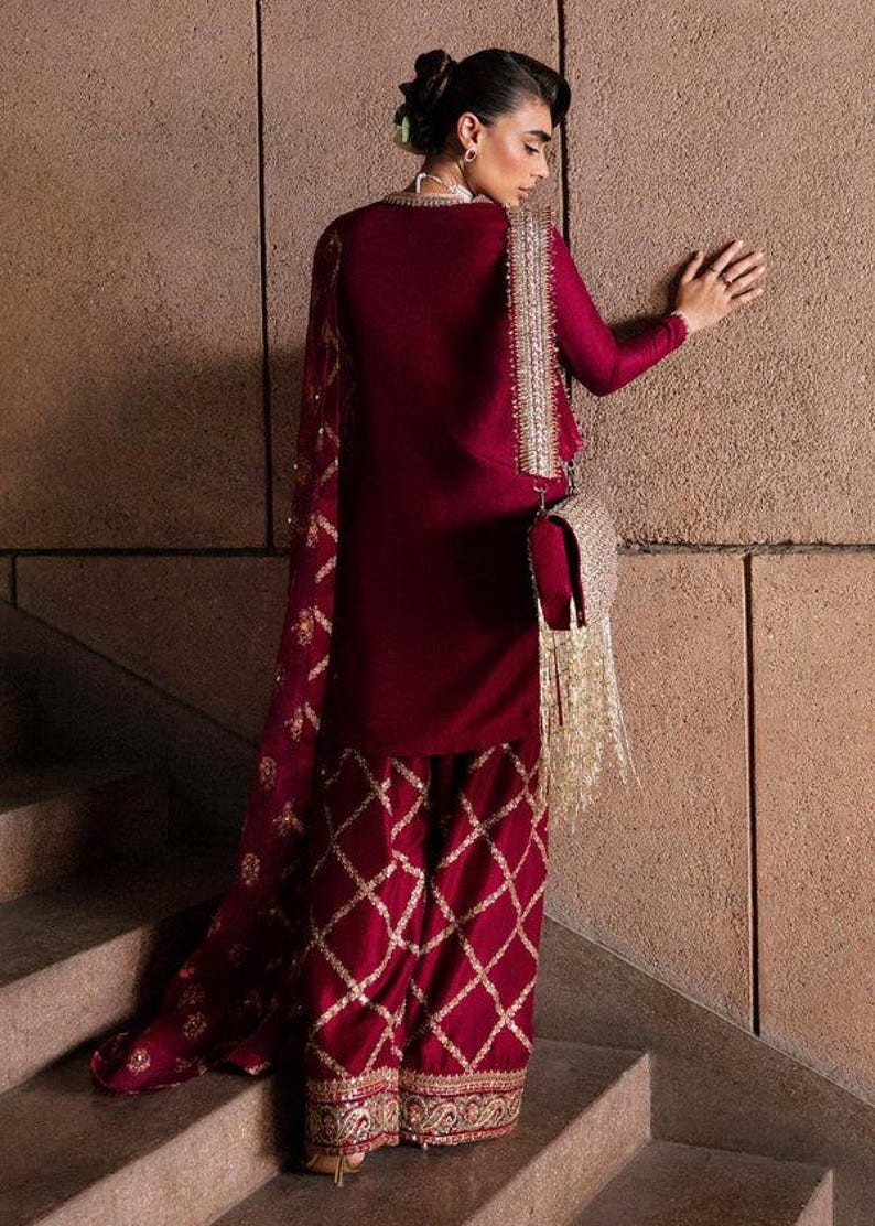 May include: A burgundy ensemble featuring a long-sleeved top and matching pants with a diamond pattern. The outfit includes a sheer scarf and a small, embellished purse with gold fringe. The pants and purse have gold accents.