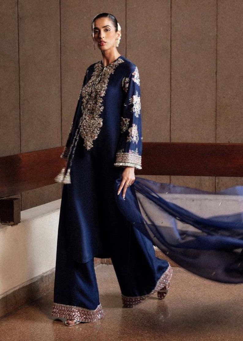 Puede incluir: Conjunto azul marino con t&uacute;nica de manga larga con bordados dorados y pantal&oacute;n de pernera ancha con ribete de lentejuelas. Una bufanda transparente ondea. La persona lleva sandalias.