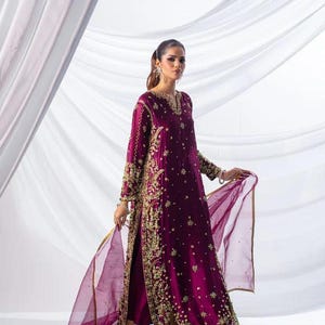 May include: A long, plum-colored formal dress with intricate gold embroidery. The dress features a high slit and long sleeves. A matching sheer scarf with gold trim is held by the wearer. The dress is paired with gold heels.