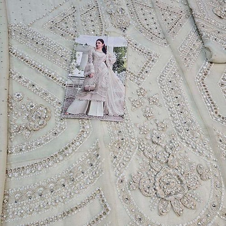 May include: Close-up of a light-colored fabric with intricate embroidery and embellishments. The fabric features a repeating pattern of floral and geometric designs, adorned with sequins and beads. A printed image of a woman in a dress is on top of the fabric.