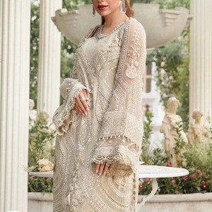 May include: An ivory-colored, long-sleeved gown with intricate embroidery and beadwork. The dress features a high neckline, flared sleeves, and a flowing skirt. The gown is embellished with delicate patterns and textures, creating an elegant and formal look.