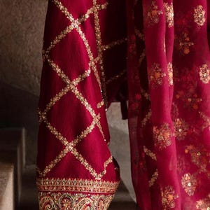 May include: Close-up of a burgundy garment with intricate gold embroidery. The fabric features a diamond pattern with gold embellishments, and the hem is adorned with detailed floral and geometric designs. The garment's flowing drape is visible.