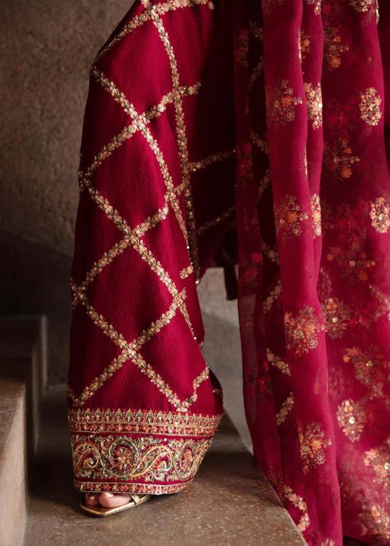 May include: Close-up of a burgundy garment with intricate gold embroidery. The fabric features a diamond pattern with gold embellishments, and the hem is adorned with detailed floral and geometric designs. The garment's flowing drape is visible.