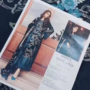 May include: A fashion magazine featuring a woman wearing a black and teal embroidered dress with a matching shawl. The magazine is on a black fabric with a white floral pattern. The text "MARIA.B. WINTER Lure" is visible.