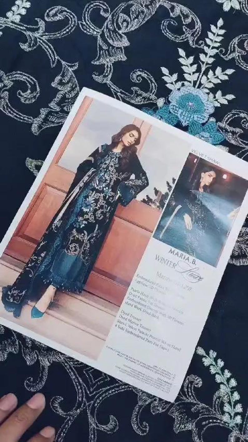 May include: A fashion magazine featuring a woman wearing a black and teal embroidered dress with a matching shawl. The magazine is on a black fabric with a white floral pattern. The text "MARIA.B. WINTER Lure" is visible.