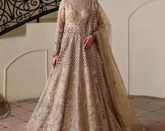 May include: A beige, floor-length formal gown with long sleeves and intricate embroidery. The dress features a fitted bodice and a flowing skirt, with a sheer shawl. The gown is embellished with sequins and beads, creating a shimmering effect. The background includes a staircase.