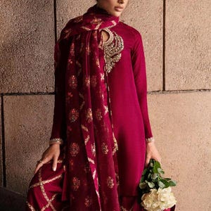 May include: A burgundy-colored ensemble featuring a long-sleeved tunic and matching wide-leg pants. The outfit includes a patterned scarf and is adorned with gold embellishments. The person is holding a bouquet of white flowers.
