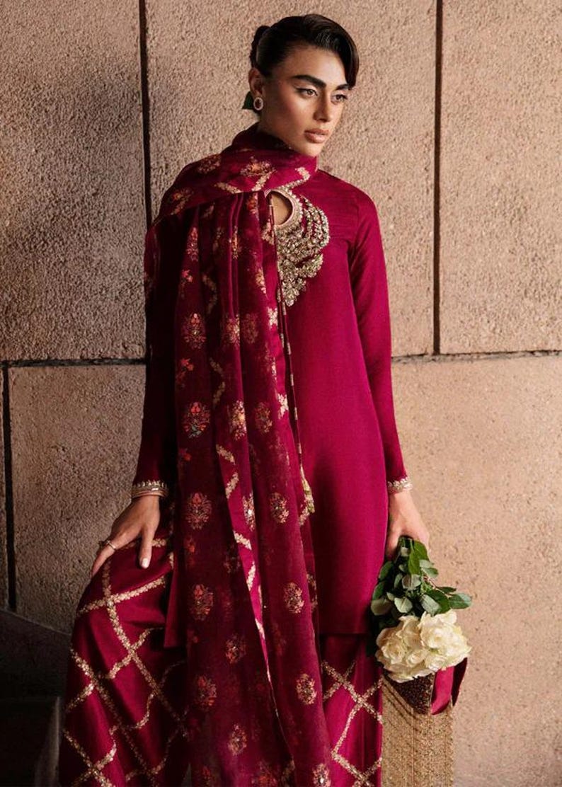May include: A burgundy-colored ensemble featuring a long-sleeved tunic and matching wide-leg pants. The outfit includes a patterned scarf and is adorned with gold embellishments. The person is holding a bouquet of white flowers.