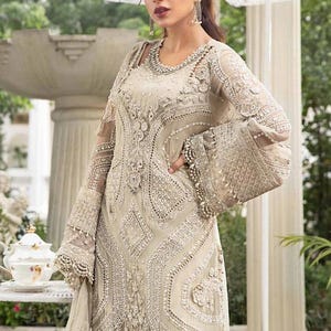 May include: An ivory-colored formal gown with intricate beadwork and embroidery. The dress features long, sheer sleeves with flared cuffs, a detailed bodice, and a flowing skirt. The gown is embellished with pearls and sequins, creating a luxurious look.