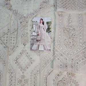 May include: A light green fabric with intricate silver beadwork and embroidery. The design features geometric patterns and floral motifs. A photograph of a woman in a matching outfit is placed on the fabric, showcasing the design and style of the garment.