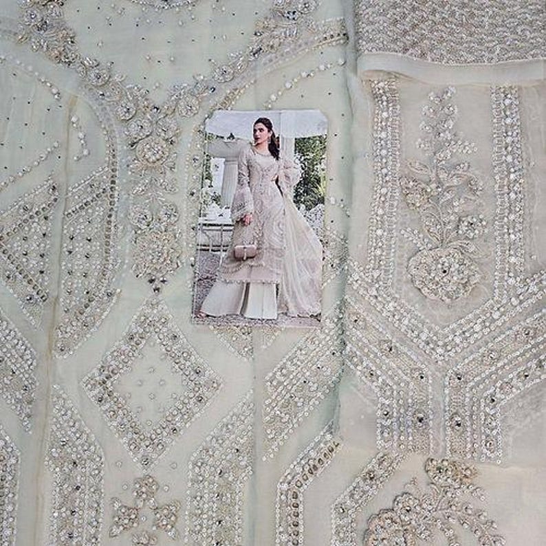 May include: A light green fabric with intricate silver beadwork and embroidery. The design features geometric patterns and floral motifs. A photograph of a woman in a matching outfit is placed on the fabric, showcasing the design and style of the garment.