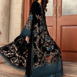 May include: A long, black velvet dress with intricate gold and teal floral embroidery. The dress features a flowing design with a teal lace trim and is paired with teal heels. The sleeves are long and flared, and the dress has a high neckline.