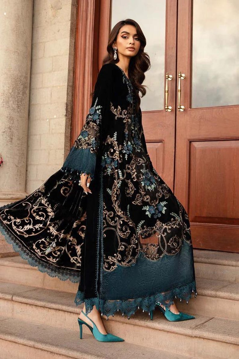 May include: A long, black velvet dress with intricate gold and teal floral embroidery. The dress features a flowing design with a teal lace trim and is paired with teal heels. The sleeves are long and flared, and the dress has a high neckline.