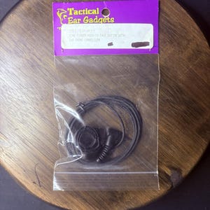 May include: A black ring finger push-to-talk button with an earphone connection, packaged in a clear plastic bag with a purple header card labeled Tactical Ear Gadgets. The text on the label reads 281/EP-PTT Ring Finger Push-To-Talk Button With Ear Phone Connection.