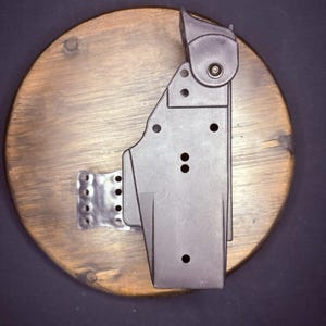 May include: A dark brown, rigid polymer holster featuring a vertical orientation with a top-mounted locking mechanism, multiple circular mounting holes along the central spine, and a metallic, perforated attachment plate extending from the side.