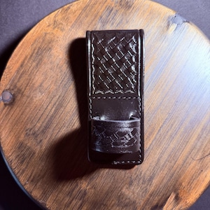 May include: A dark brown leather sheath featuring a rectangular body with a basketweave pattern and a smaller circular loop attached to the front, also embossed with a matching basketweave texture, secured with visible dark brown stitching along the edges.