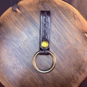 May include: A dark brown leather keychain strap featuring a vertical embossed sunburst pattern along the center. The strap is secured with a circular brass-toned metal rivet and holds a matching brass-toned metal ring at the bottom end.