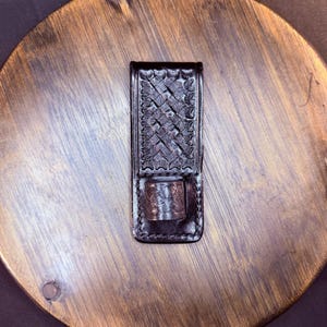 May include: A dark brown leather sheath featuring a rectangular body with a basket-weave embossed pattern and a small, cylindrical leather loop attached to the lower front section, secured with visible stitching along the edges of the leather.