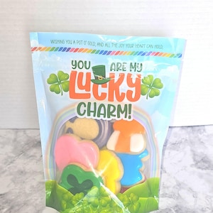 May include: A clear cellophane bag filled with decorated sugar cookies. The bag has a blue background with a rainbow and the words "You Are My Lucky Charm!" The cookies are shaped like shamrocks, hearts, and leprechauns.