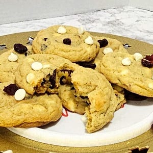 May include: A plate of freshly baked cookies with white chocolate chips and dried cranberries. The cookies are on a gold-colored plate with decorative cutouts. One cookie is broken, revealing the interior.