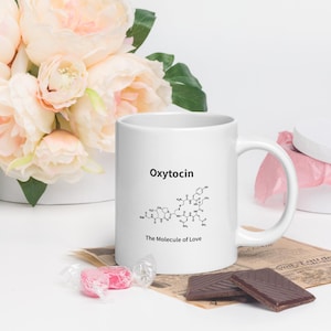 May include: White ceramic mug with the word "Oxytocin" and a molecular structure graphic. Below the graphic, the text reads "The Molecule of Love". The mug is next to a bouquet of pink flowers and chocolate.