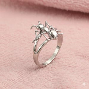 May include: A silver ring with an ant motif. The ant is the focal point, with its body and legs forming the ring's main design. The ring has a split band, adding a modern twist to the insect-inspired jewellery. The ring is made of a shiny metal.