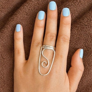May include: A silver ring with a unique design featuring a rectangular frame and an interior swirl. The ring is worn on a hand with light blue nail polish. The background is a brown, textured surface.