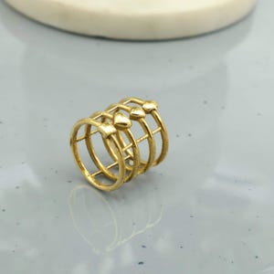Op de afbeelding: Een goudkleurige ring met een uniek ontwerp. De ring heeft drie banden die zijn verbonden door verticale staven en versierd met hartvormige accenten. De ring wordt gepresenteerd op een lichtgrijze ondergrond.