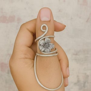 May include: A silver-toned ring with a floral design, wrapped around a finger. The ring features a flower in the center and a curved design that wraps around the finger. The ring is a piece of jewelry.