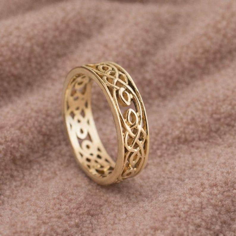 May include: A gold-colored ring with an intricate Celtic knot design. The ring features a continuous pattern of interwoven lines and open spaces, creating a detailed and textured appearance. The ring is displayed on a neutral, textured surface.